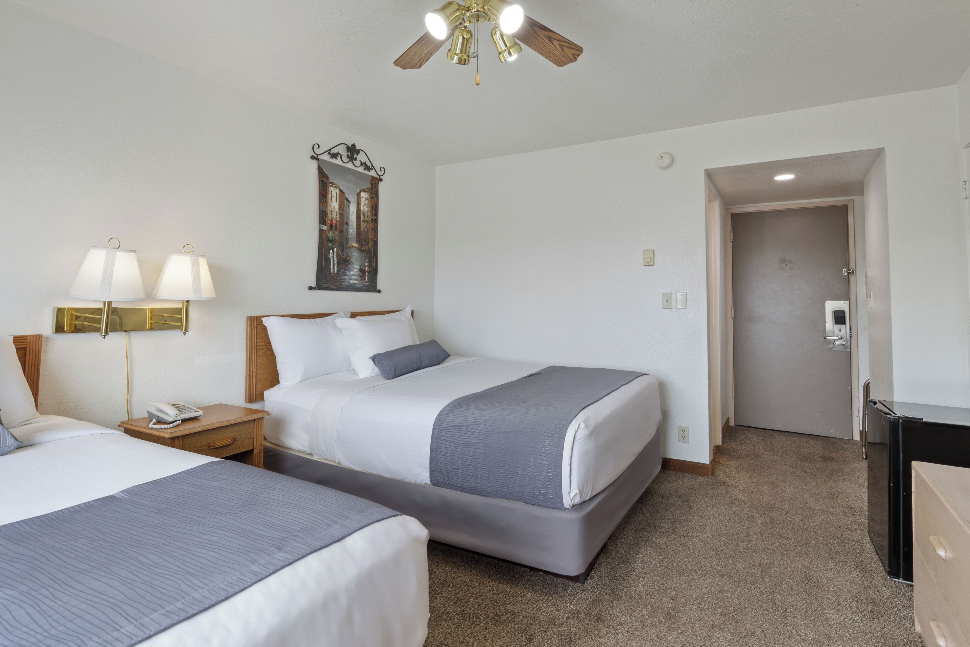 Habitación de hotel con dos camas, un ventilador de techo, un cuadro, un teléfono en la mesita de noche y una puerta que da a otra habitación.