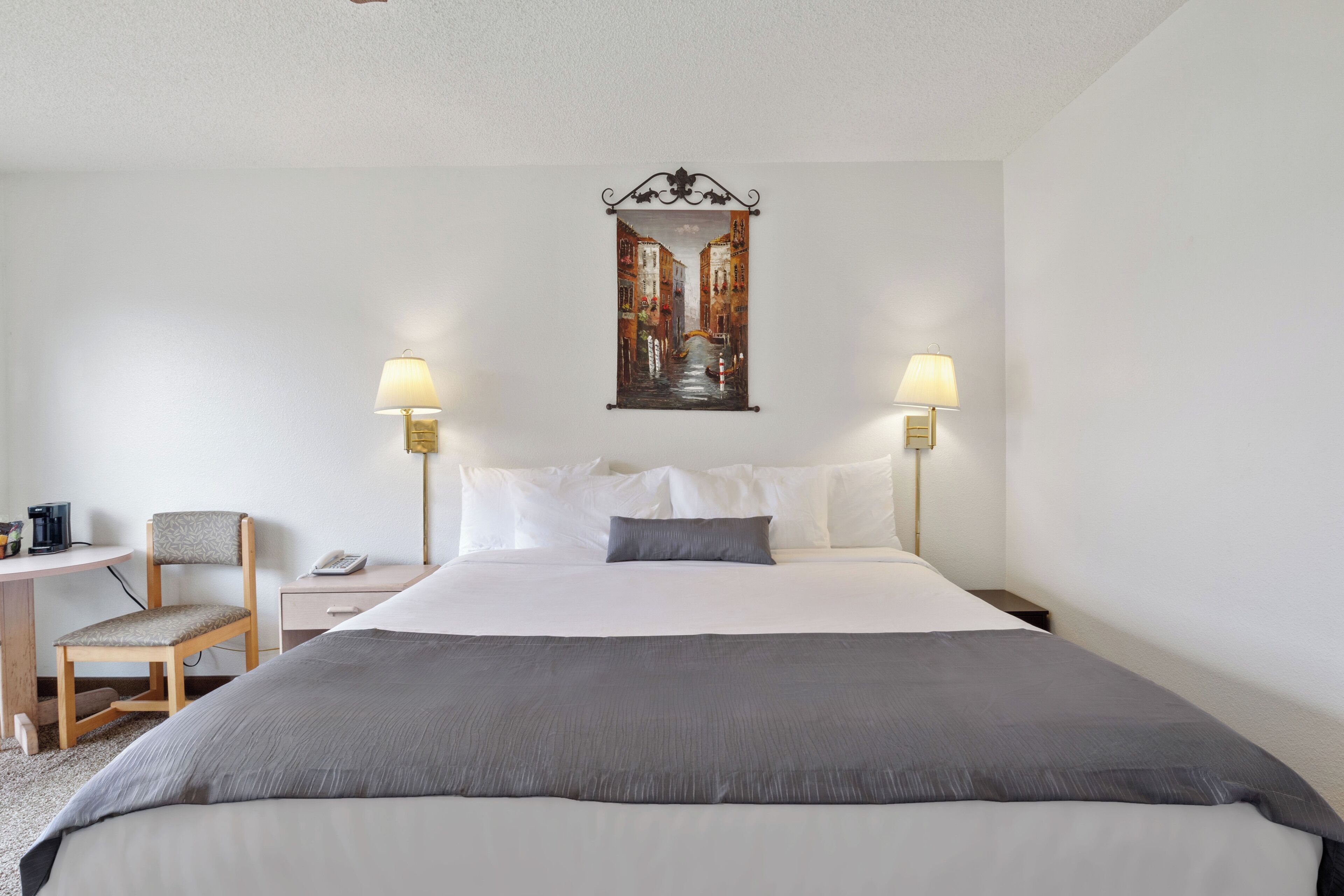 A hotel room with a bed, two bedside lamps, a chair, a small table, and a painting on the wall.