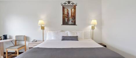 A hotel room with a bed, two bedside lamps, a chair, a small table, and a painting on the wall.