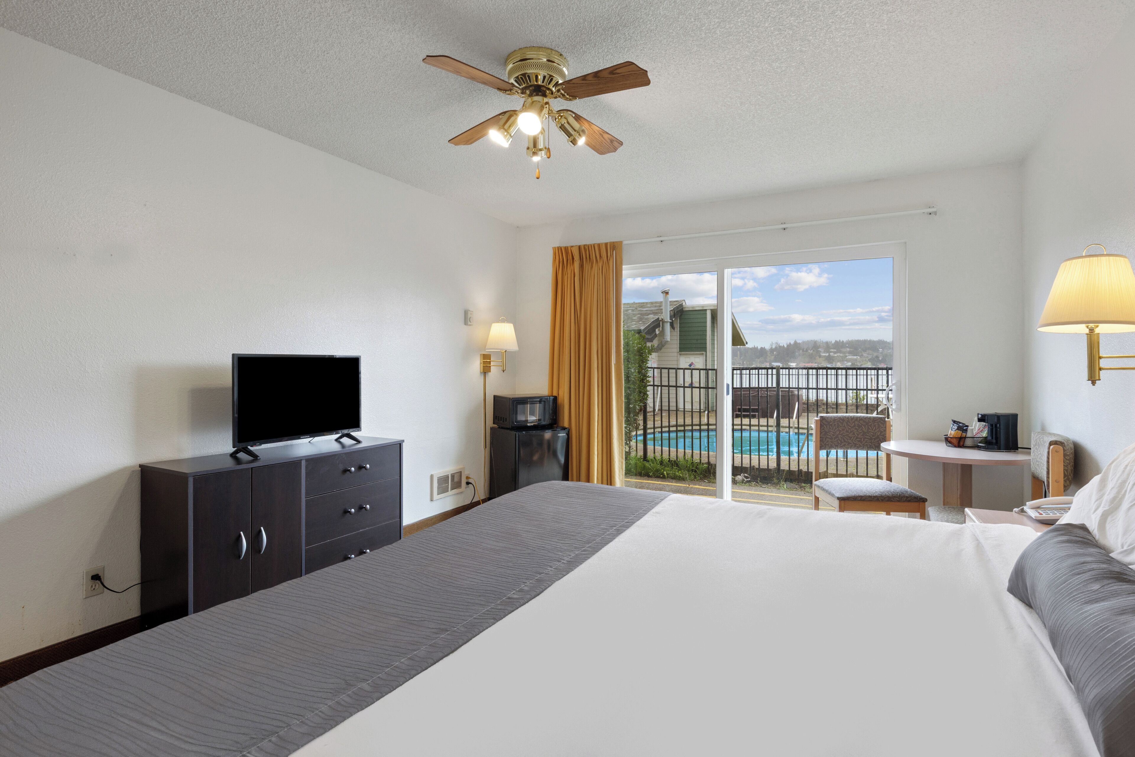 A hotel room with a bed, a television, a desk with a lamp, a chair, and a view of a swimming pool.