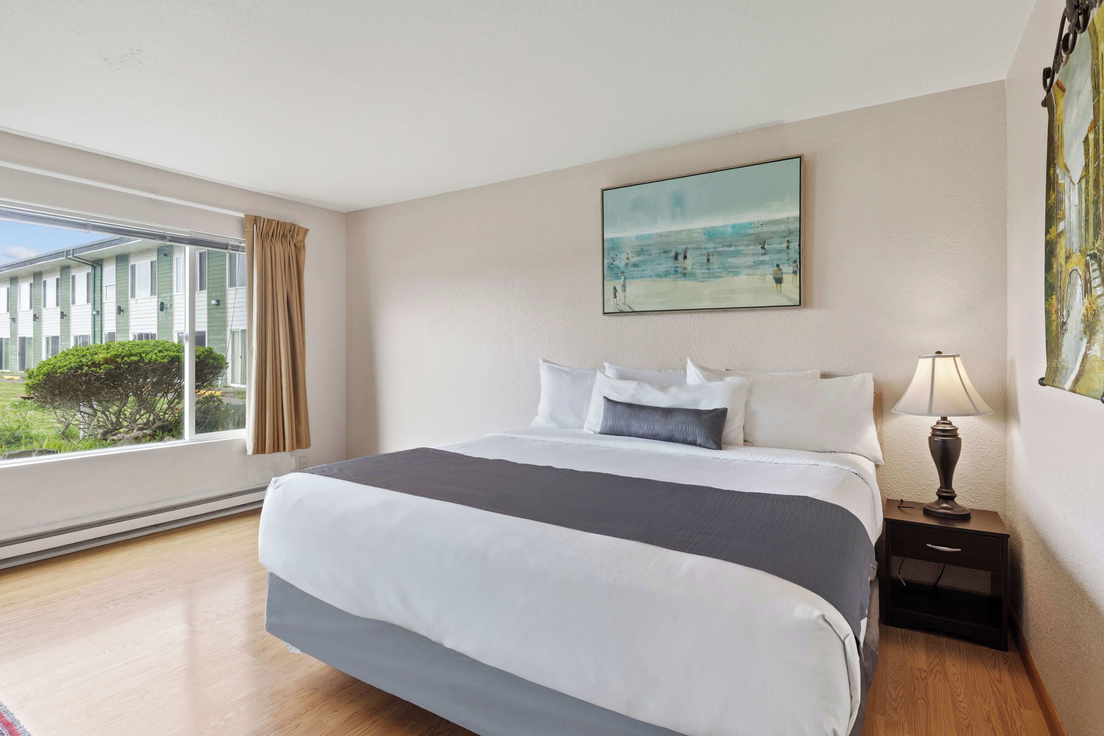 Habitación de hotel con una cama grande, una mesita de noche con lámpara, un cuadro con una playa y vista a un edificio a través de la ventana.