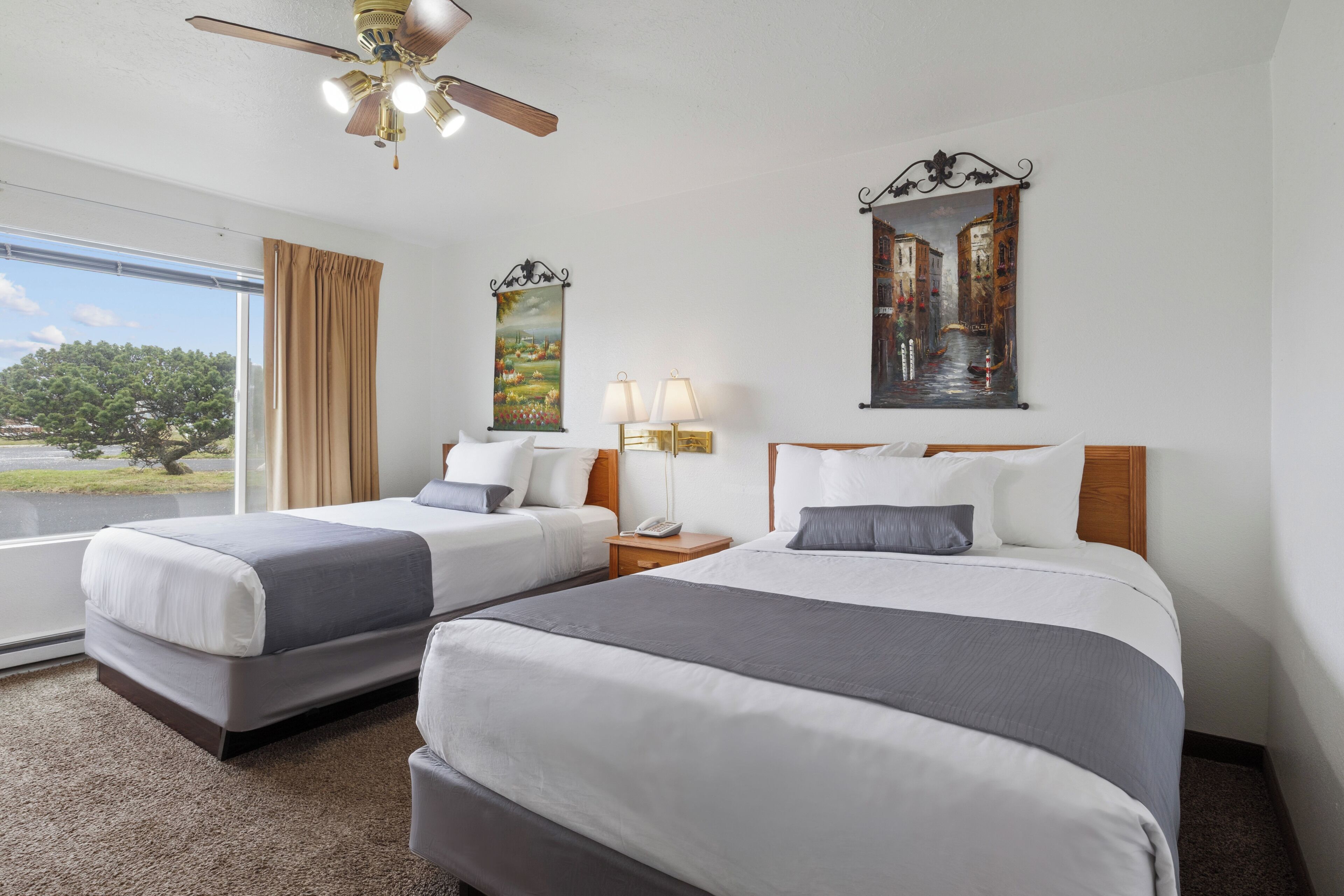 A hotel room with two beds, a ceiling fan, a window with curtains, a painting, and a nightstand with a lamp.