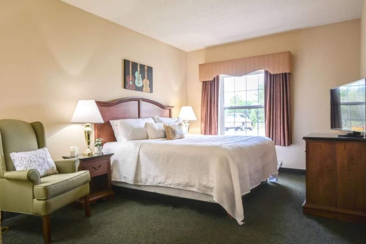 Une chambre d’hôtel avec un lit, une chaise, une table de chevet, une lampe, un téléviseur et une fenêtre avec des rideaux.