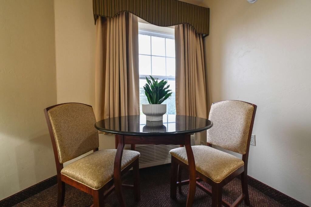 A room with a round glass table, two upholstered chairs, beige curtains, a potted plant, and a window with a view.