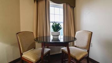 A room with a round glass table, two upholstered chairs, beige curtains, a potted plant, and a window with a view.
