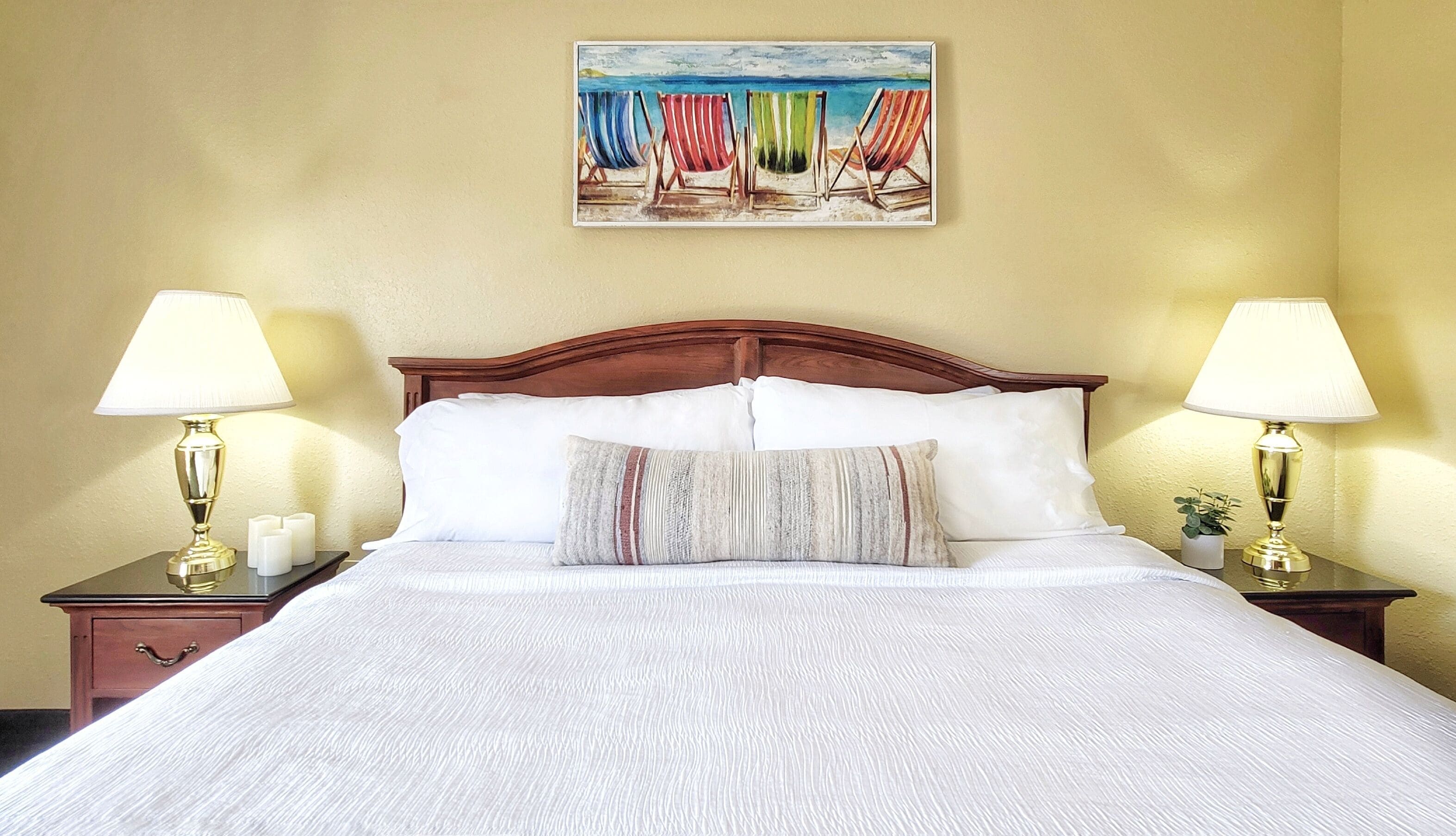 A neatly made bed with white linens, two bedside lamps, a small bedside table with a candle, and a painting of beach chairs on the wall.