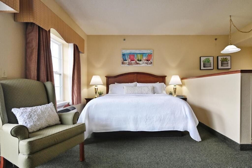 Een hotelkamer met een groot bed, twee bedlampen, een fauteuil met een kussen, een raam met gordijnen en een schilderij aan de muur.