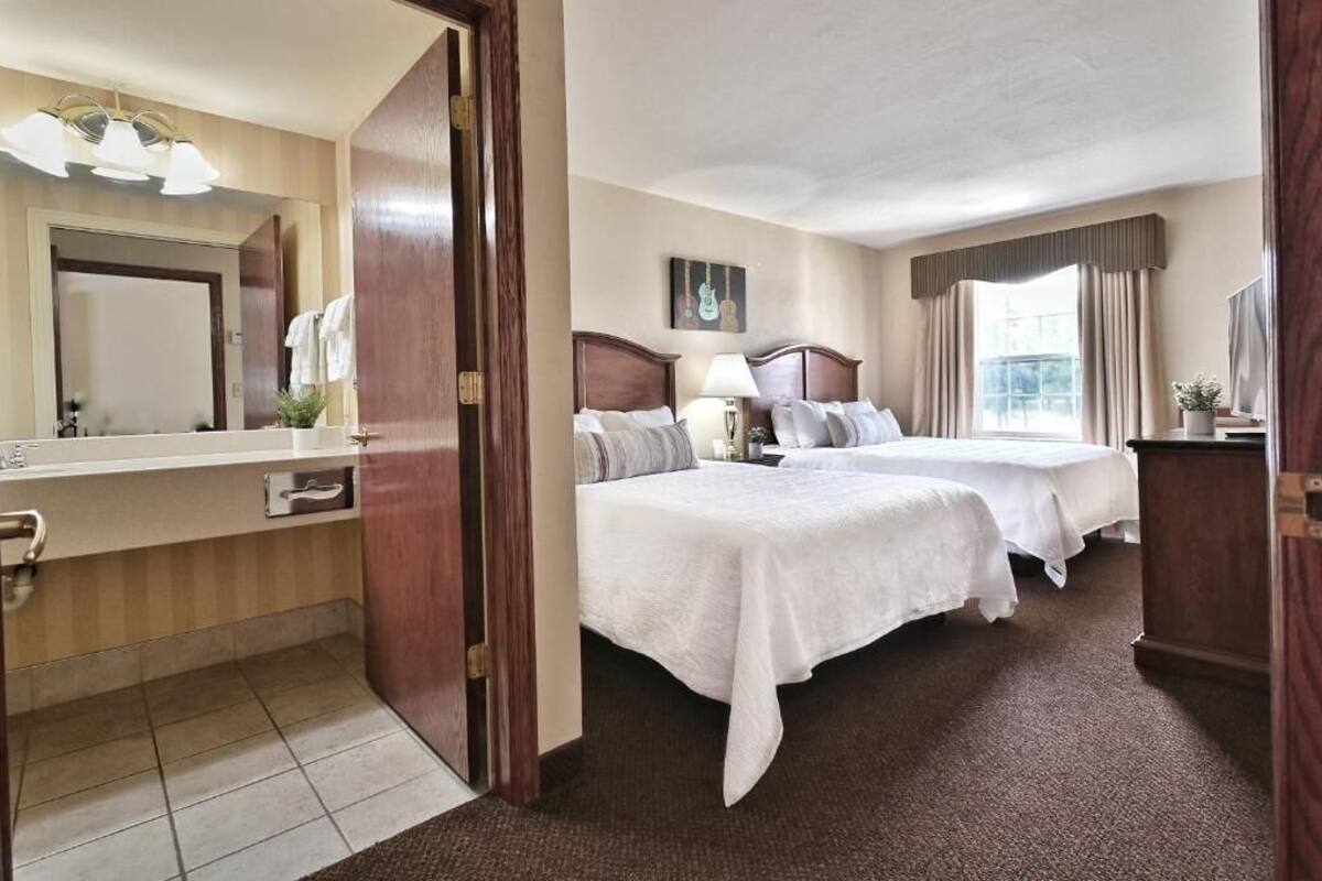 Une chambre d’hôtel avec deux lits, une salle de bain avec un miroir et un lavabo, et une fenêtre avec des rideaux.