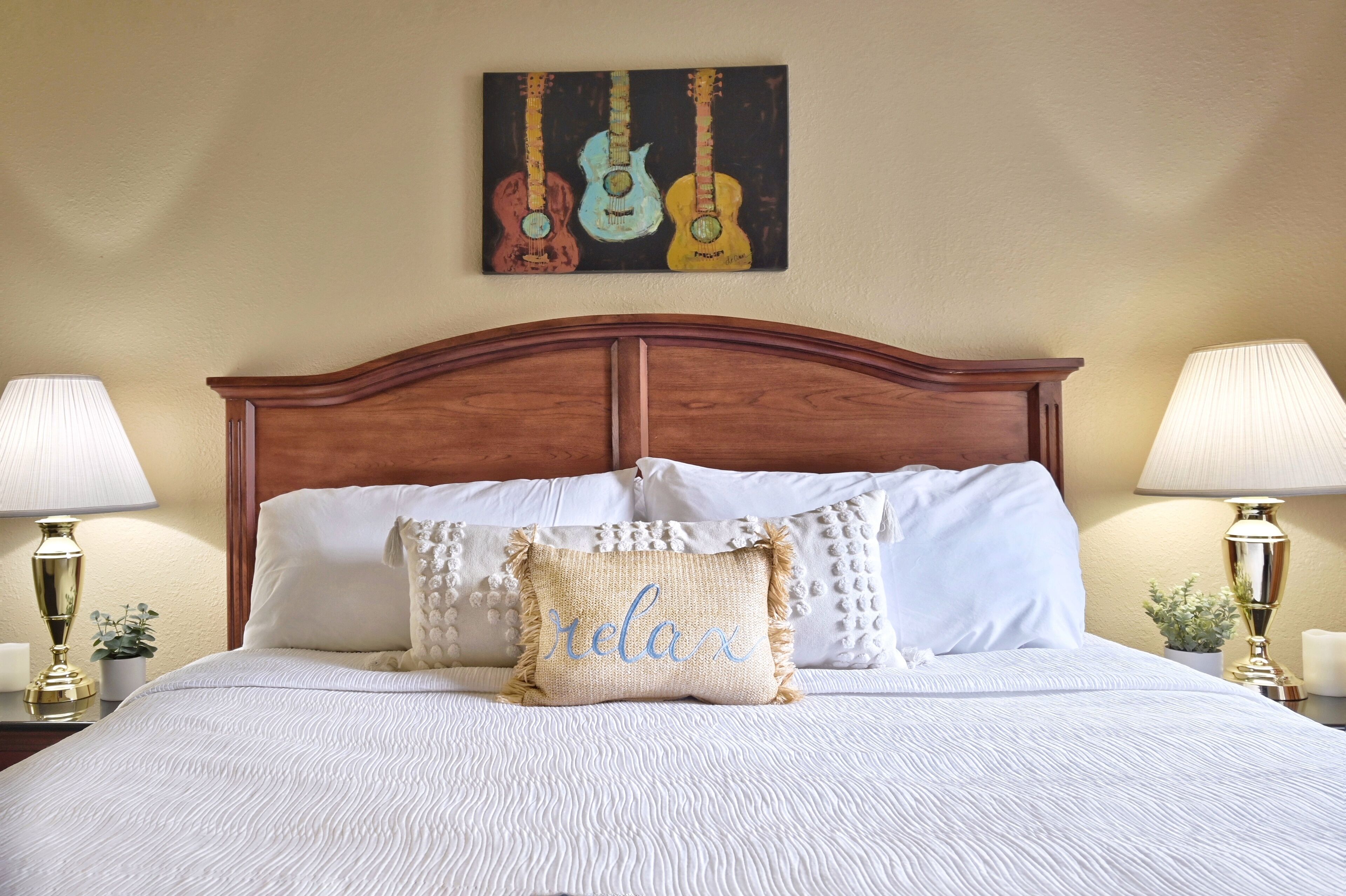 A neatly made bed with white linens, a 'relax' pillow, and a wall art of guitars.