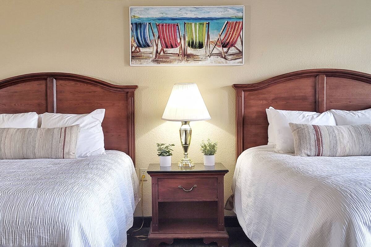 Une chambre d’hôtel avec deux lits, une table de chevet avec une lampe et un tableau représentant des chaises de plage.