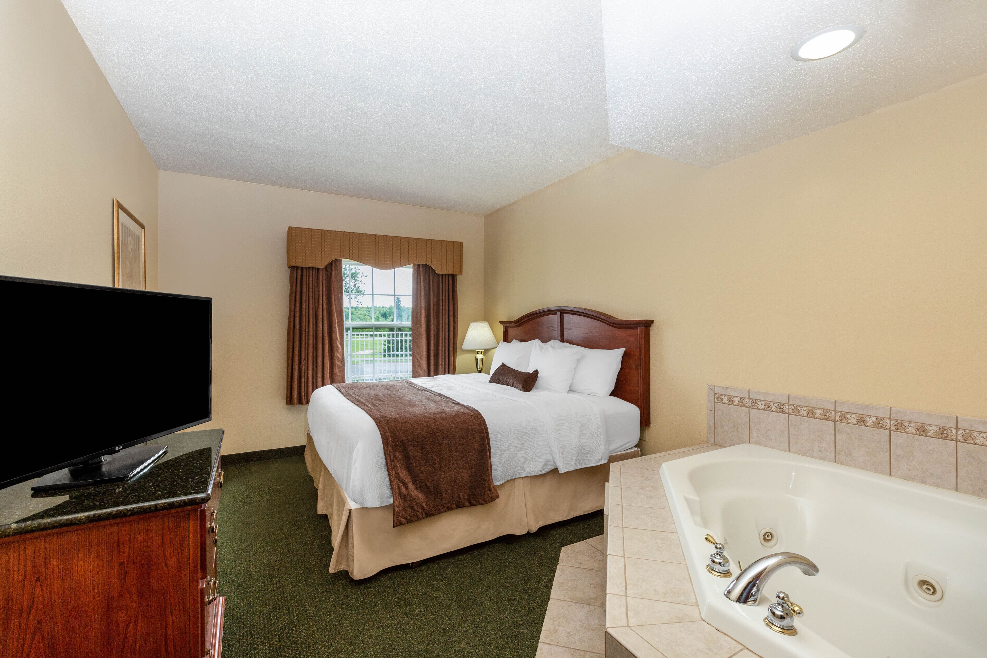 Quarto de hotel com cama, televisão, banheiro com banheira e janela com cortinas.