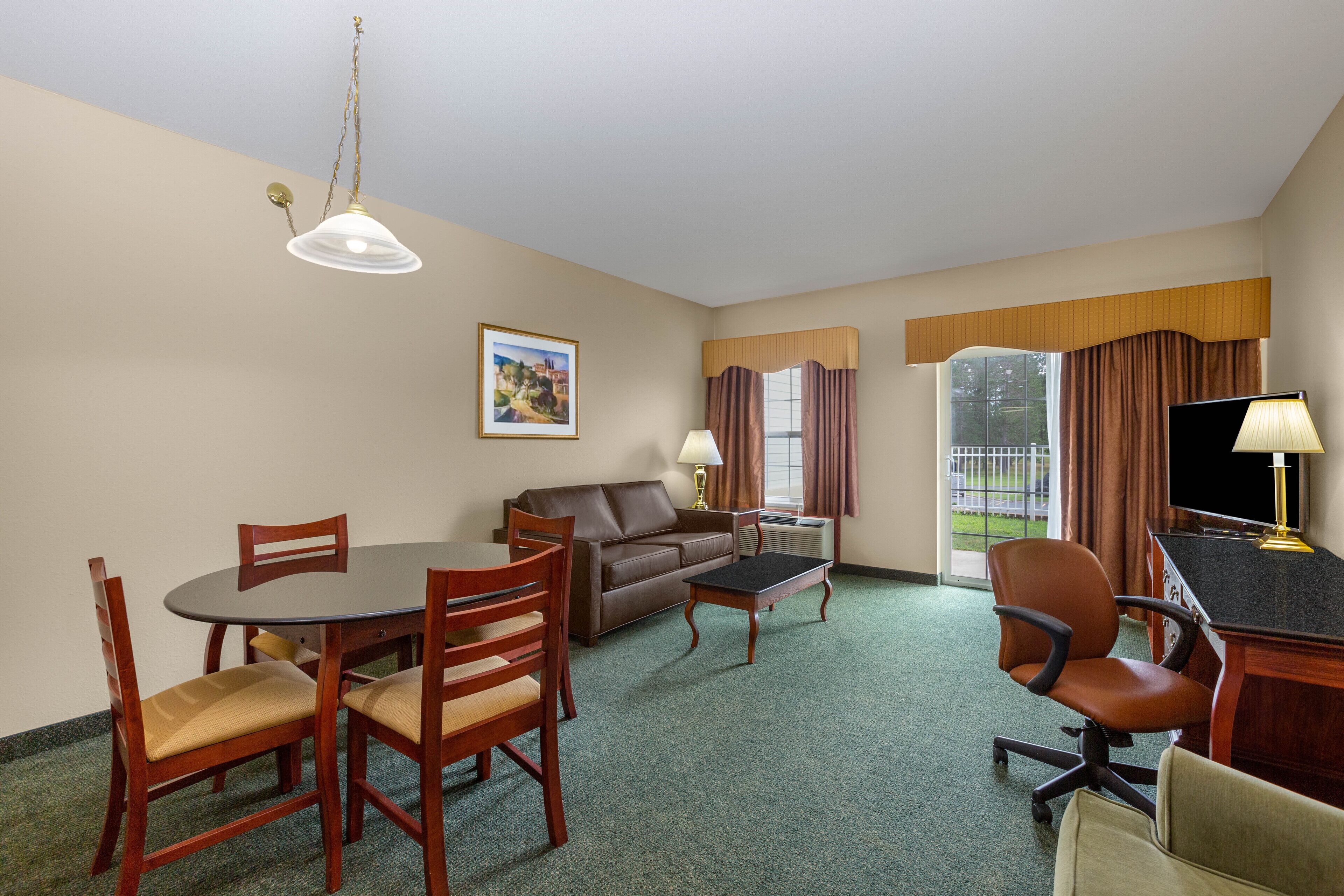 Habitación de hotel con una mesa de comedor, sillas, un sofá, un televisor y una ventana con cortinas.