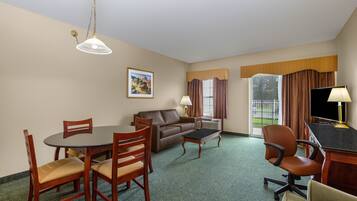 Habitación de hotel con una mesa de comedor, sillas, un sofá, un televisor y una ventana con cortinas.