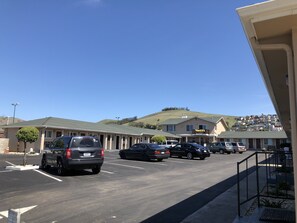 A parking lot with several cars, a building with a green roof, and a hillside in the background. -  SurfSide Stay Morro Bay (Morro Bay)