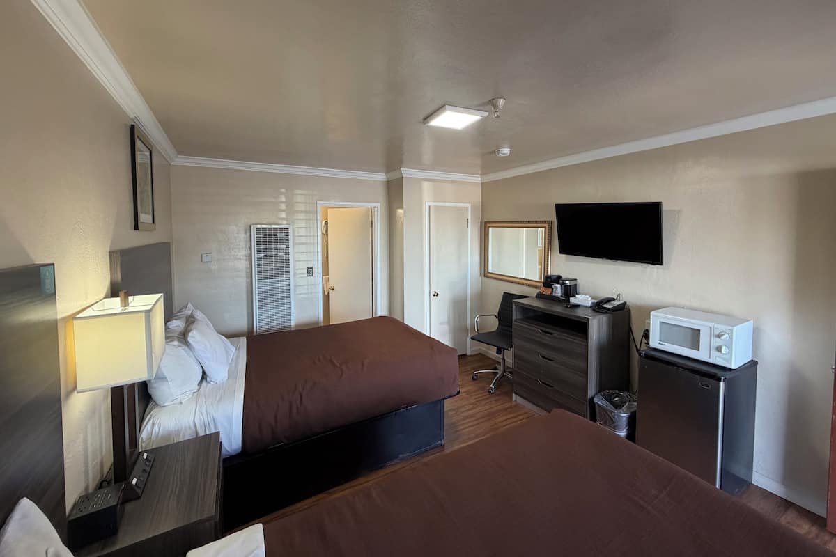 A hotel room with two beds, a desk with a lamp, a dresser, a TV, a microwave, and a ceiling fan.