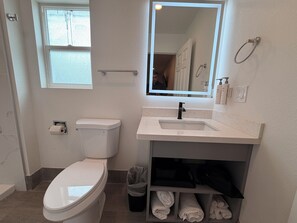 A bathroom with a toilet, sink, mirror, and towel storage. -  SurfSide Stay Morro Bay (Morro Bay)