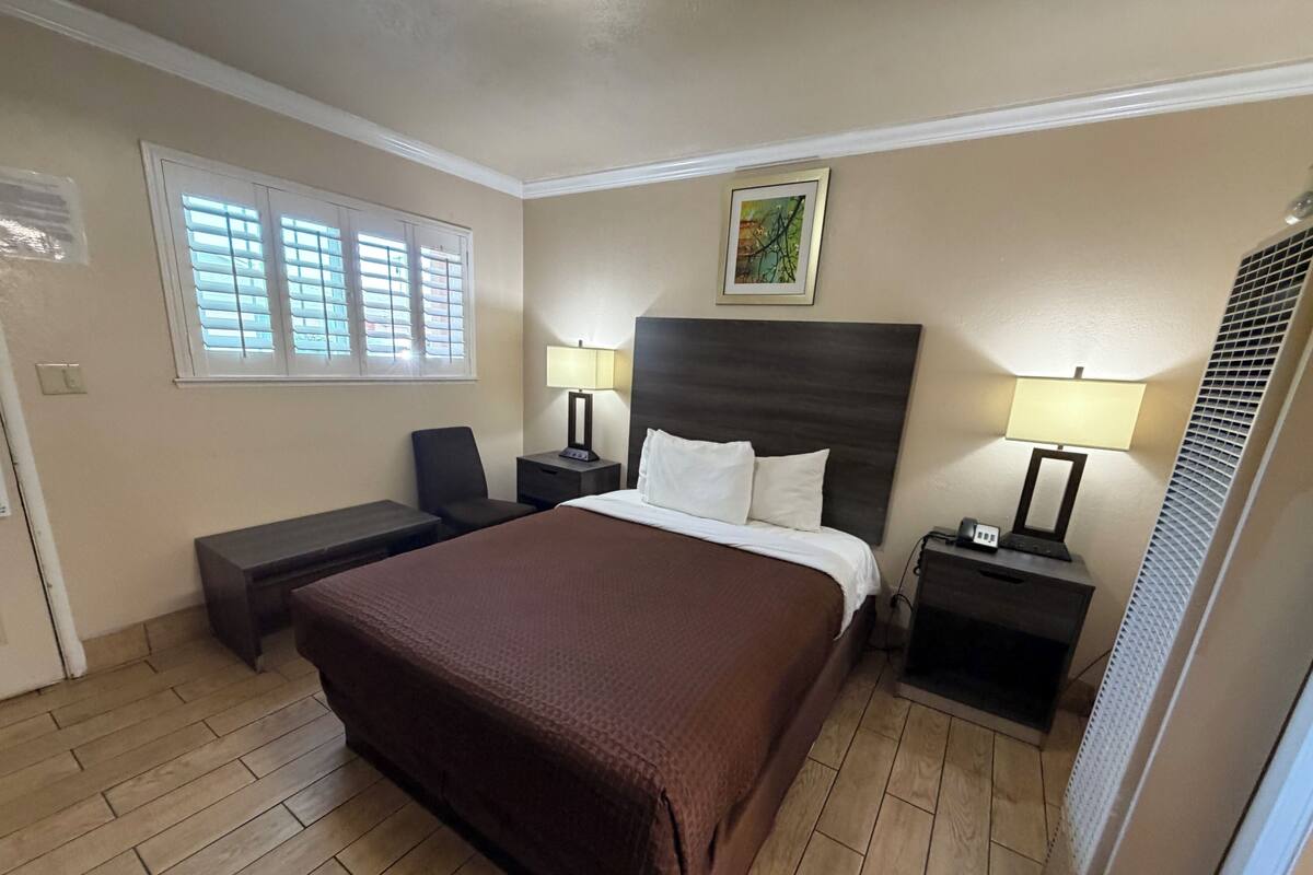 A hotel room with a large bed, two bedside tables with lamps, a chair, a desk, and a window with blinds.