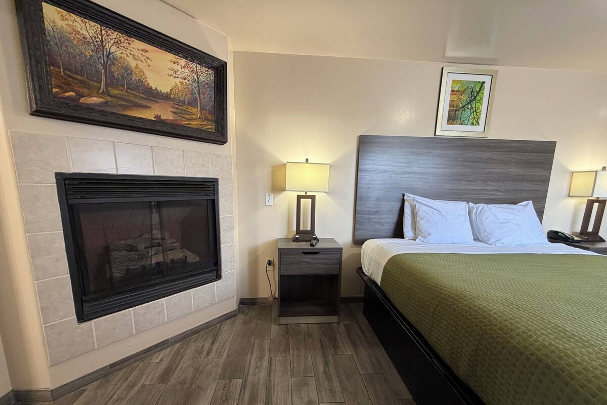 A hotel room with a fireplace, a bed with a green bedspread, two bedside lamps, and two framed pictures on the wall.