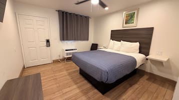A hotel room with a bed, a ceiling fan, a window with curtains, a bedside table, and a wall-mounted artwork.
