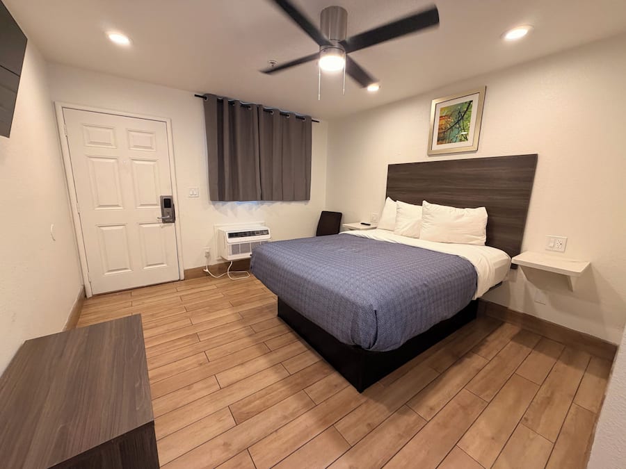 Een hotelkamer met een bed, een plafondventilator, een raam met gordijnen, een nachtkastje en een aan de muur bevestigd kunstwerk.