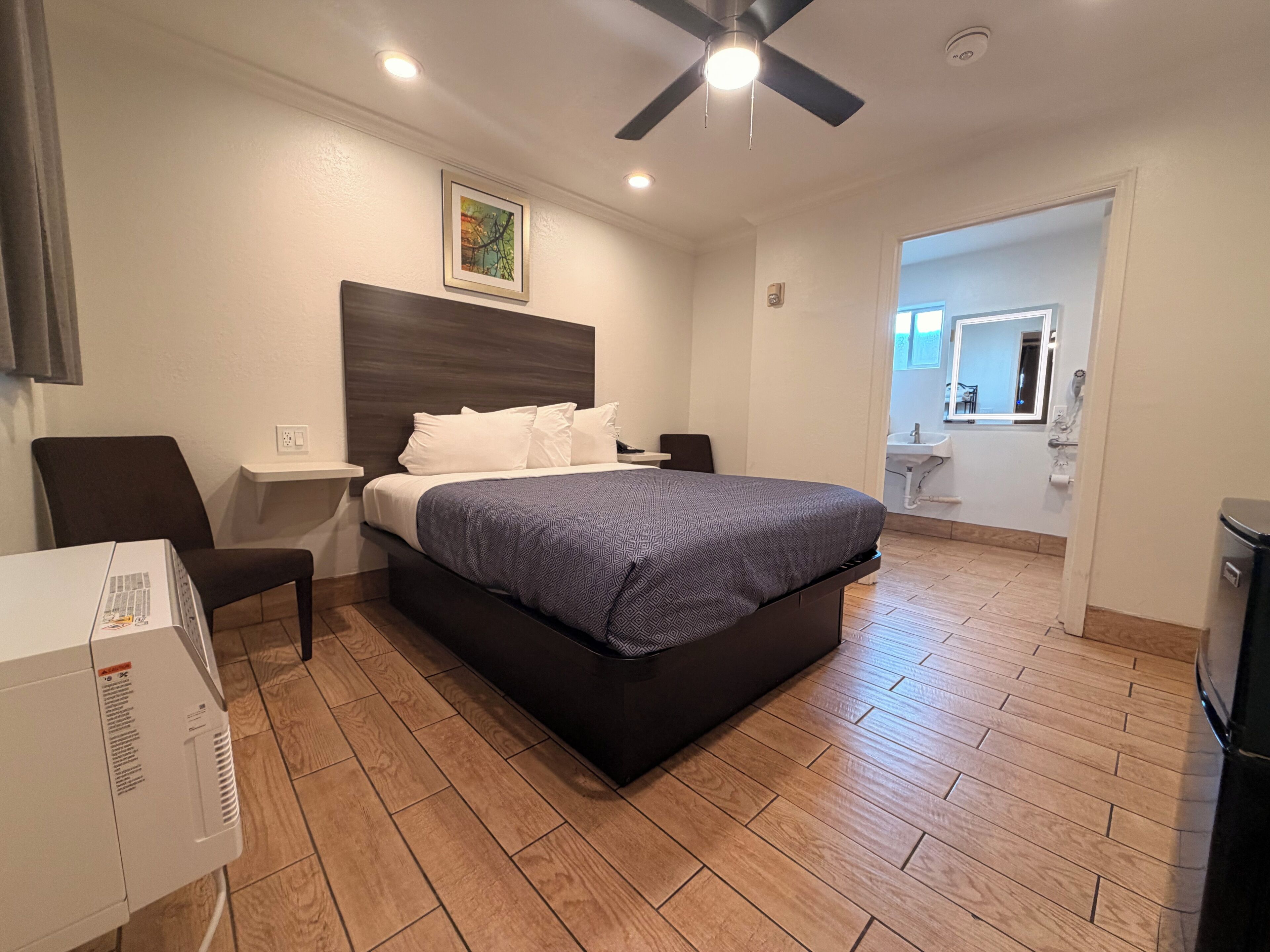 A hotel room with a bed, a desk, a chair, a ceiling fan, and a bathroom visible through an open door.