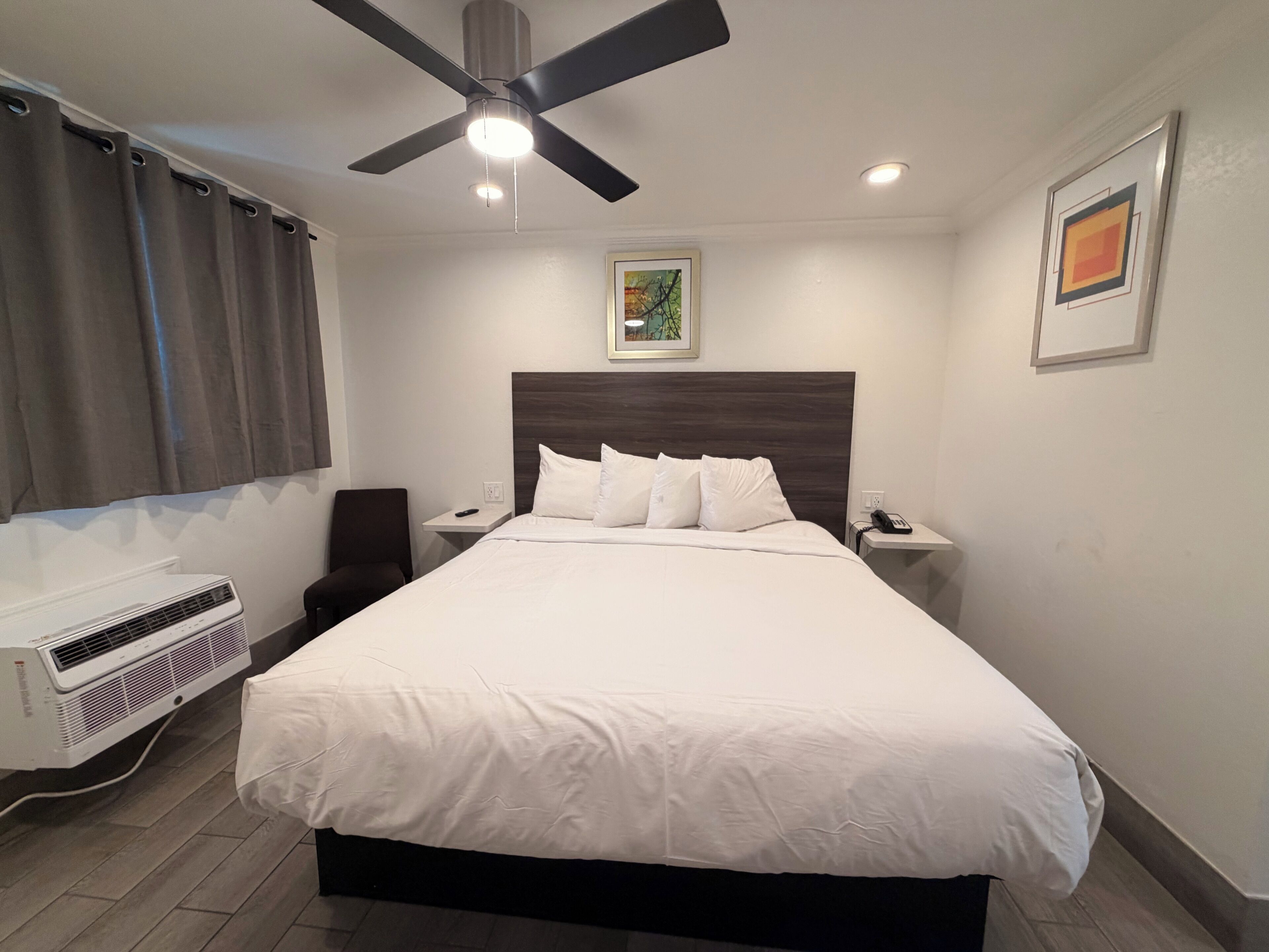 A hotel room with a bed, a ceiling fan, a bedside table with a phone, a wall-mounted air conditioner, and a painting on the wall.