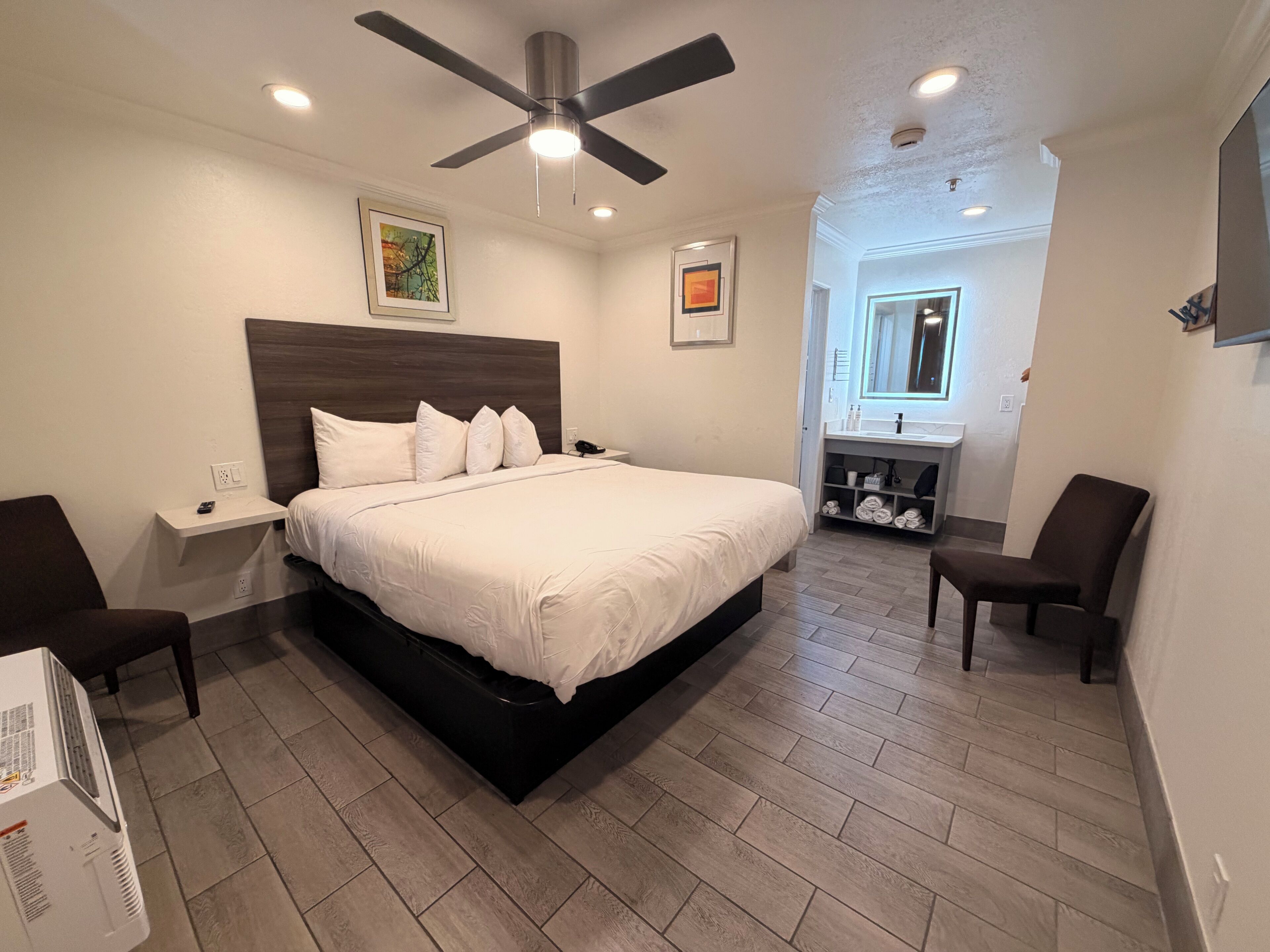 A bedroom with a bed, a nightstand, a chair, a ceiling fan, and a bathroom visible in the background.