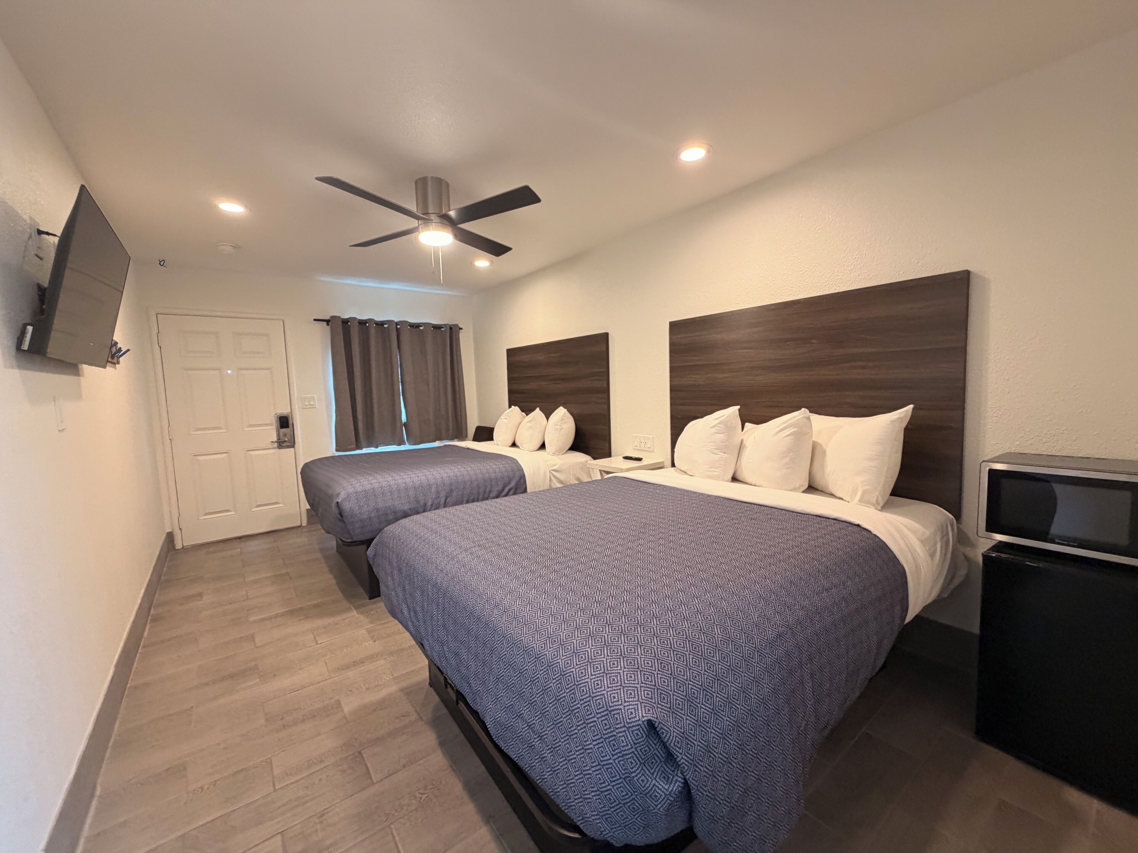 Habitación de hotel con dos camas, ventilador de techo, televisor y una puerta con cortinas.