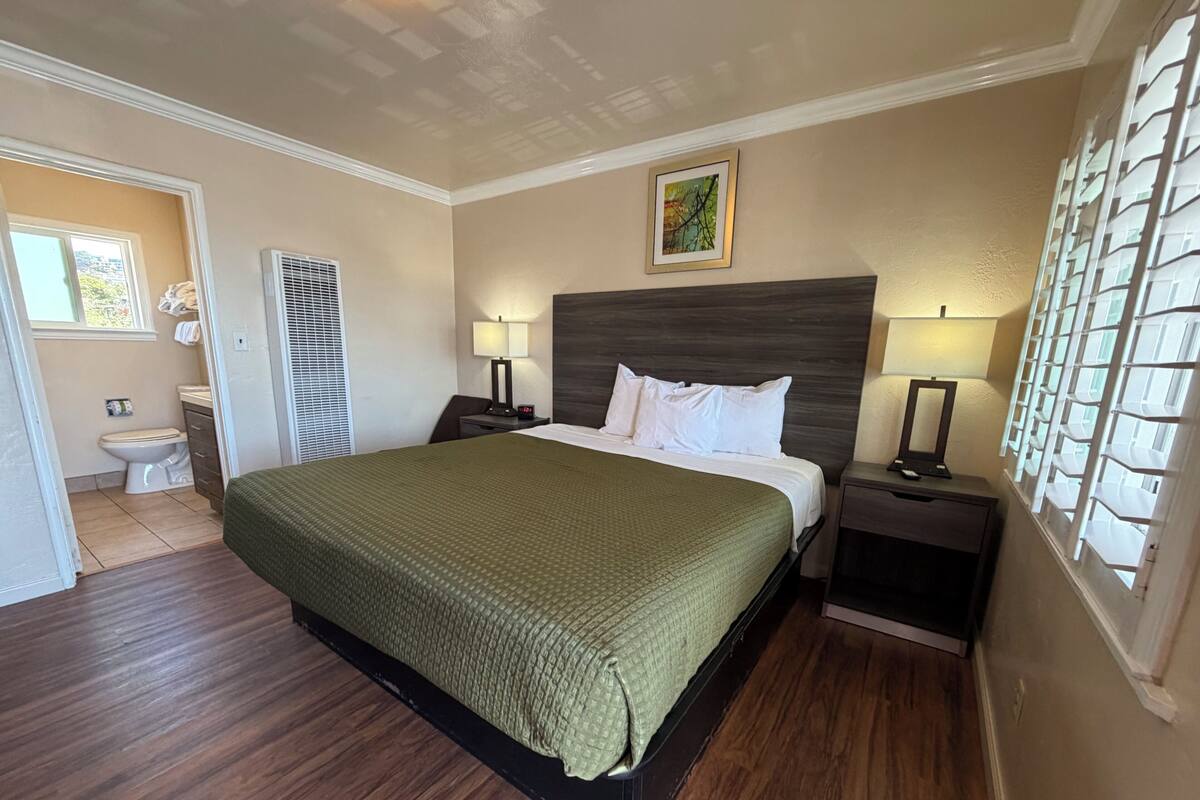 A bedroom with a bed, two bedside tables, a headboard, a window with blinds, a door leading to a bathroom, and a painting on the wall.