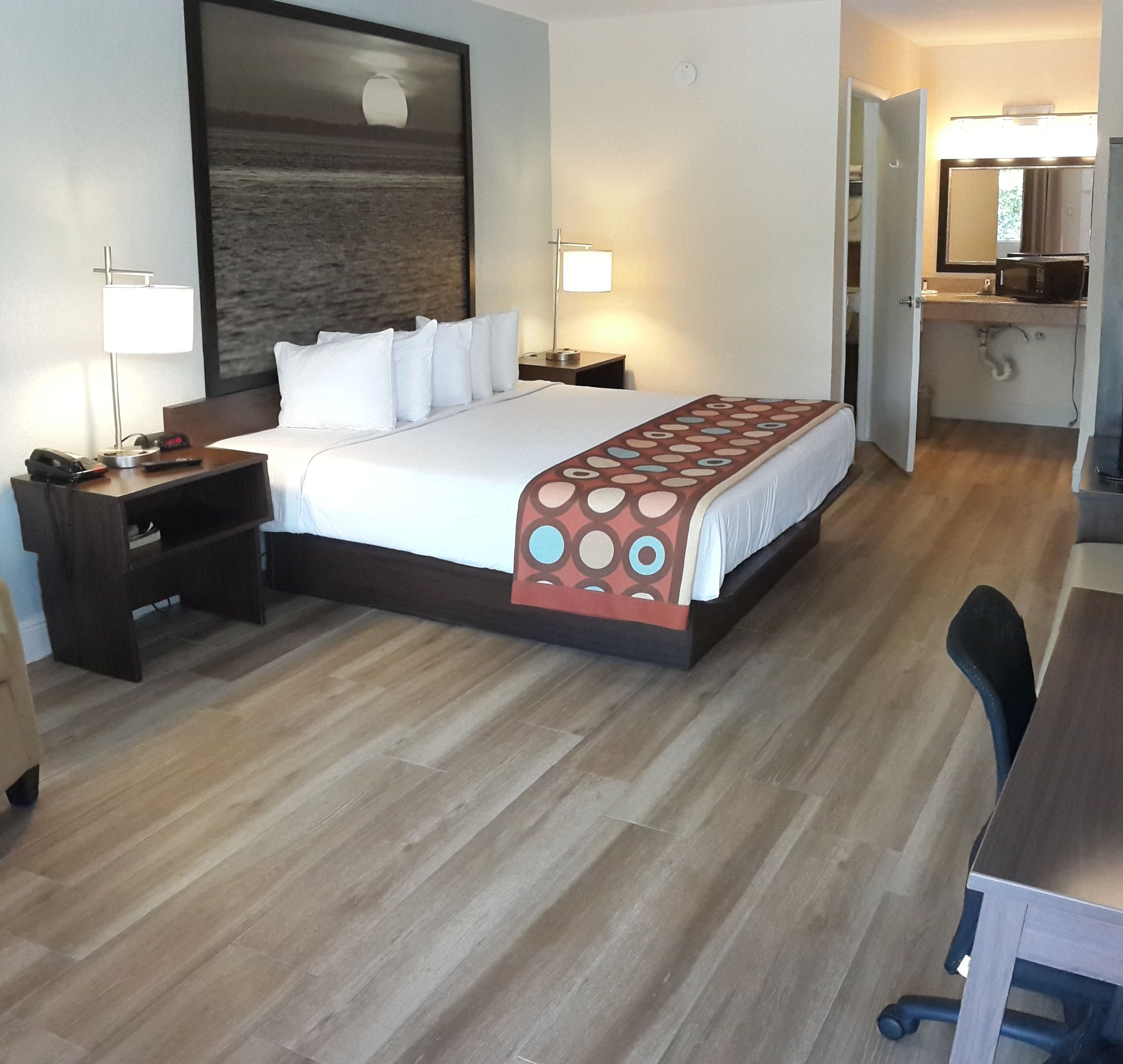 A hotel room with a bed, bedside tables, a desk, a chair, a lamp, and a large window with a view of the sea.