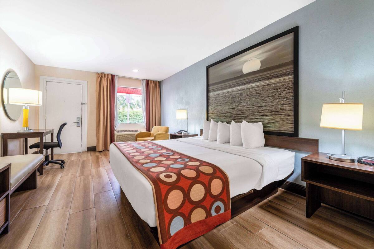 A hotel room with a large bed, a desk, a chair, a desk lamp, a bedside table, a nightstand, a wall-mounted TV, and a window with curtains.