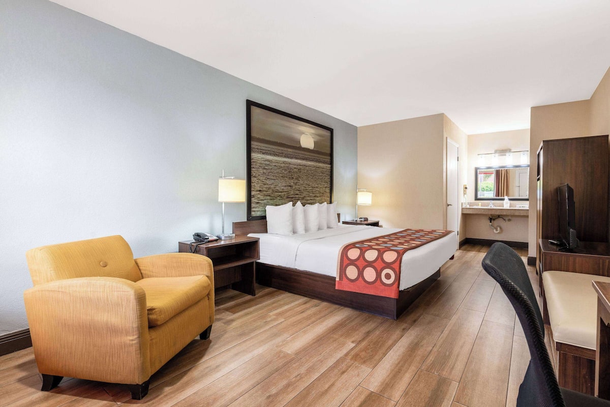 A hotel room with a bed, a yellow armchair, a desk with a lamp, a TV, and a view of the kitchen area.