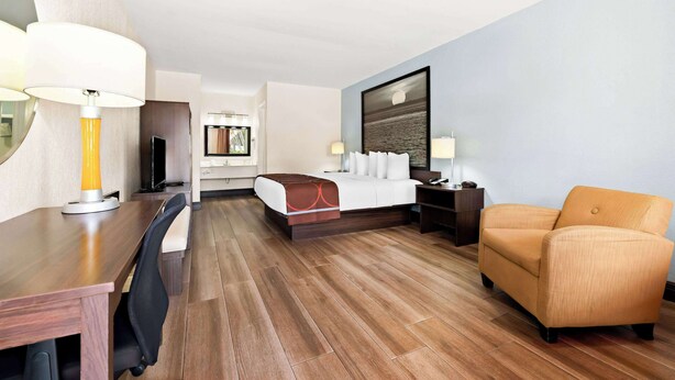 A hotel room with a wooden floor, a bed with white linens, a sofa, a desk with a lamp, and a television.