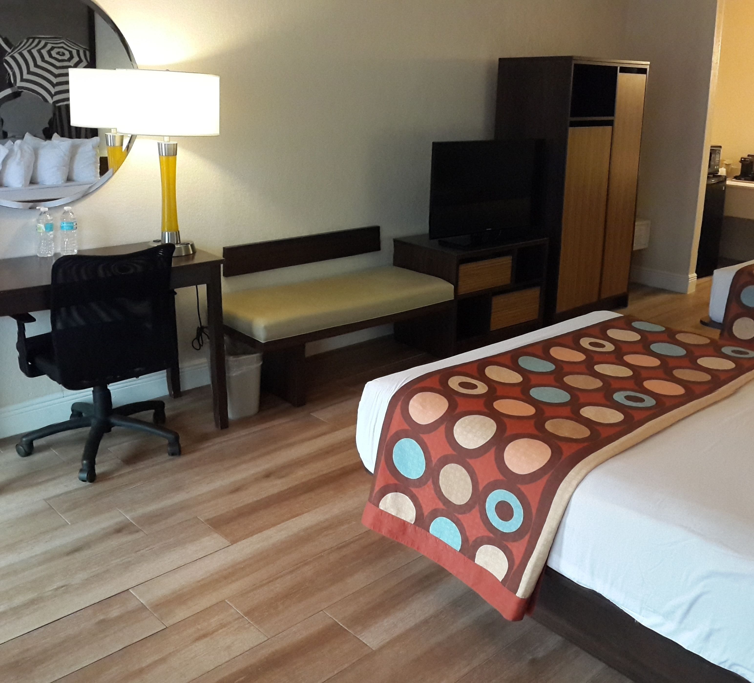 A hotel room with a bed, a desk with a chair, a television, a wardrobe, and a bench.