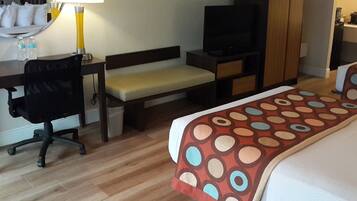 A hotel room with a bed, a desk with a chair, a television, a wardrobe, and a bench.