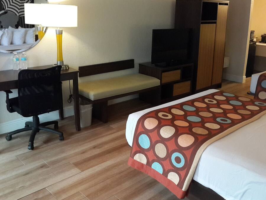 A hotel room with a bed, a desk with a chair, a television, a wardrobe, and a bench.