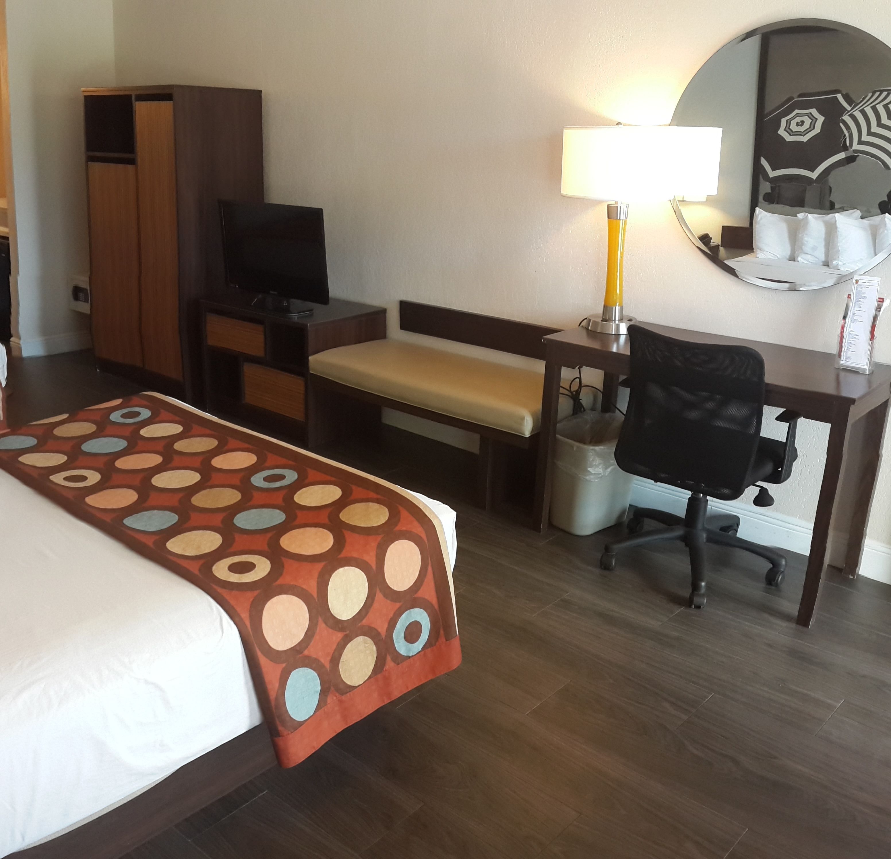 Habitación de hotel con cama, escritorio, silla, televisor, lámpara y espejo.