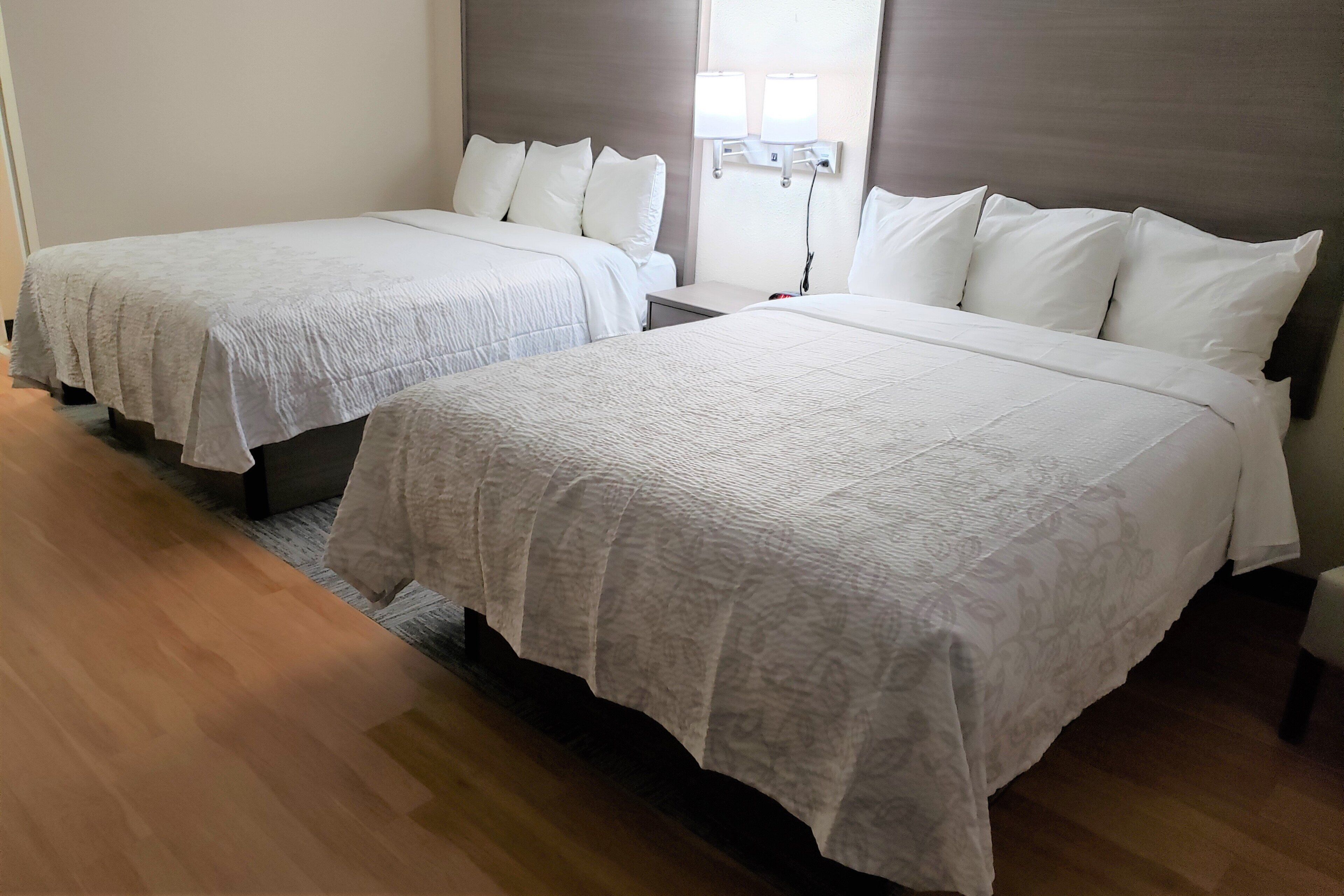 A hotel room with two beds, each with white linens and pillows, a bedside table, and a wall-mounted lamp.