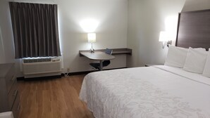 Et hotelværelse med seng, natbord, lampe, klimaanlæg og et vindue med gardiner.