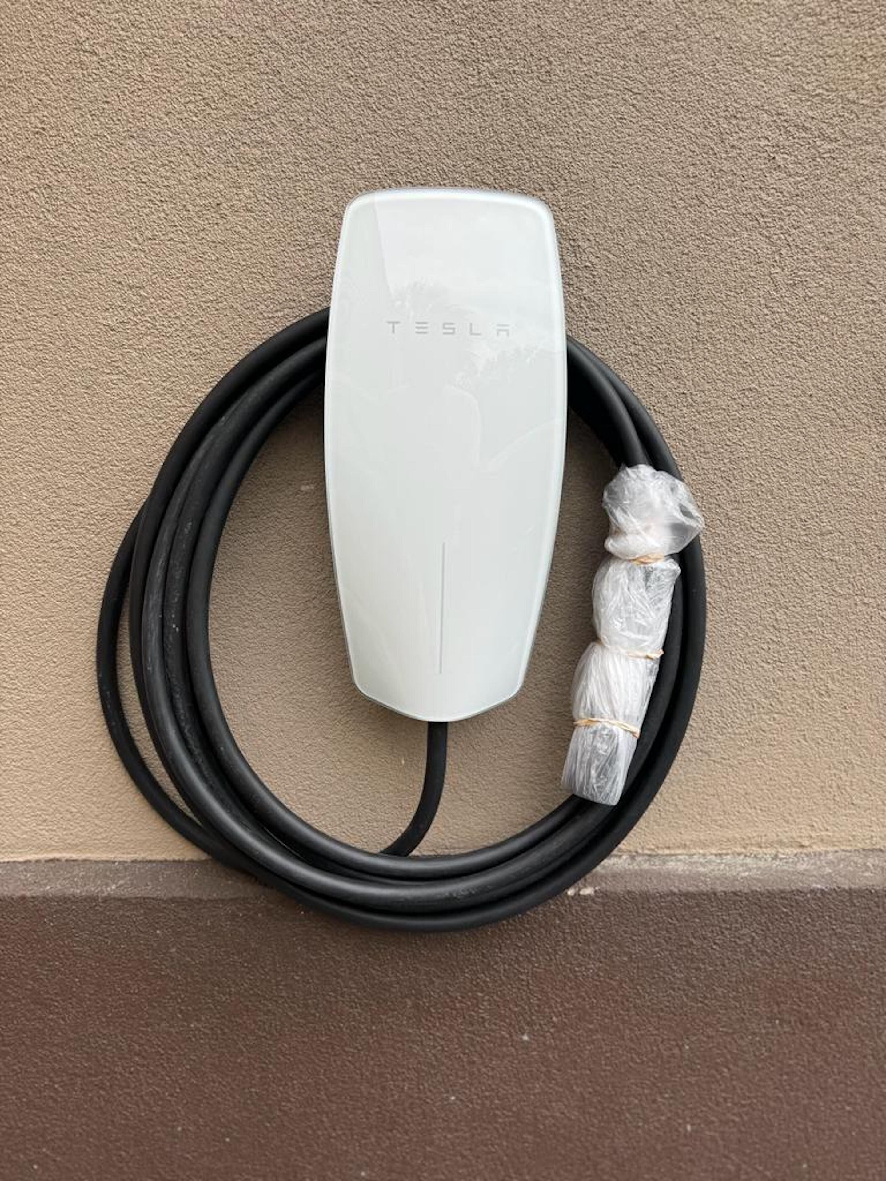 A white Tesla charging station with a black cable and plastic-wrapped cables.