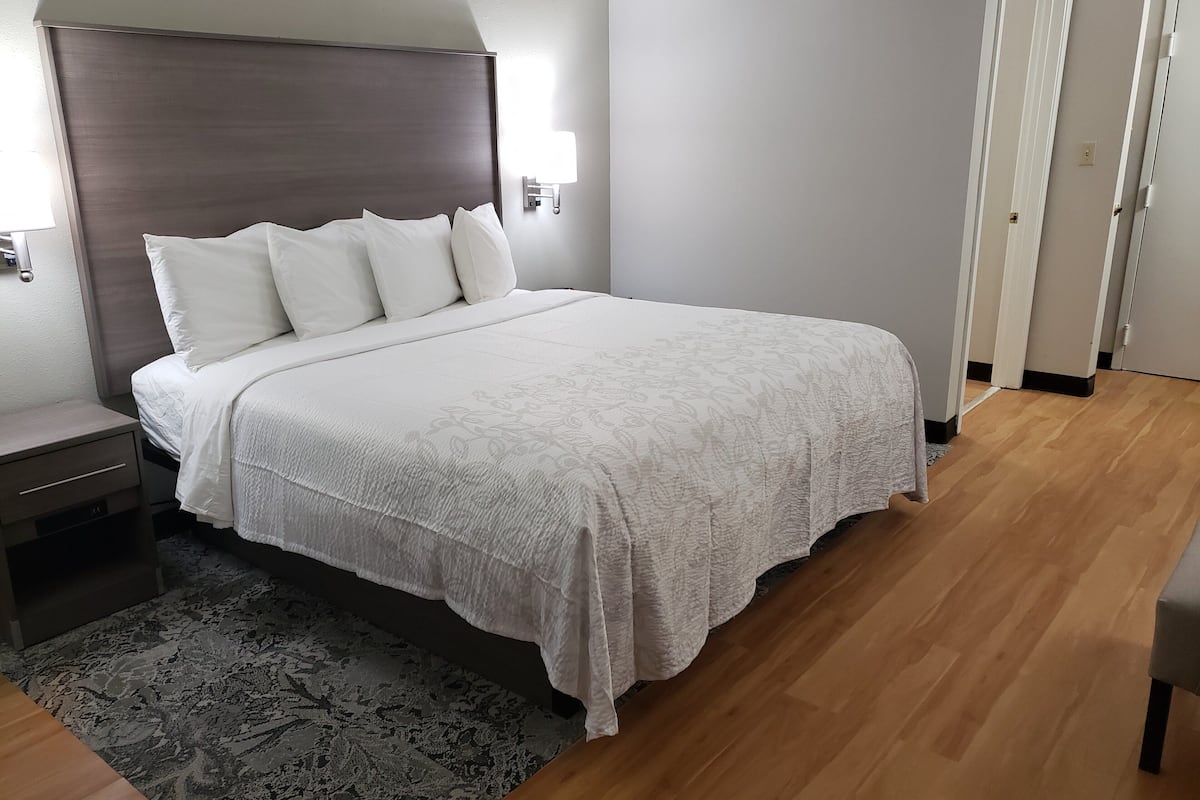 Siististi pedattu sänky valkoisilla lakanoiden kanssa, yöpöytä ja seinään kiinnitetty valaisin.