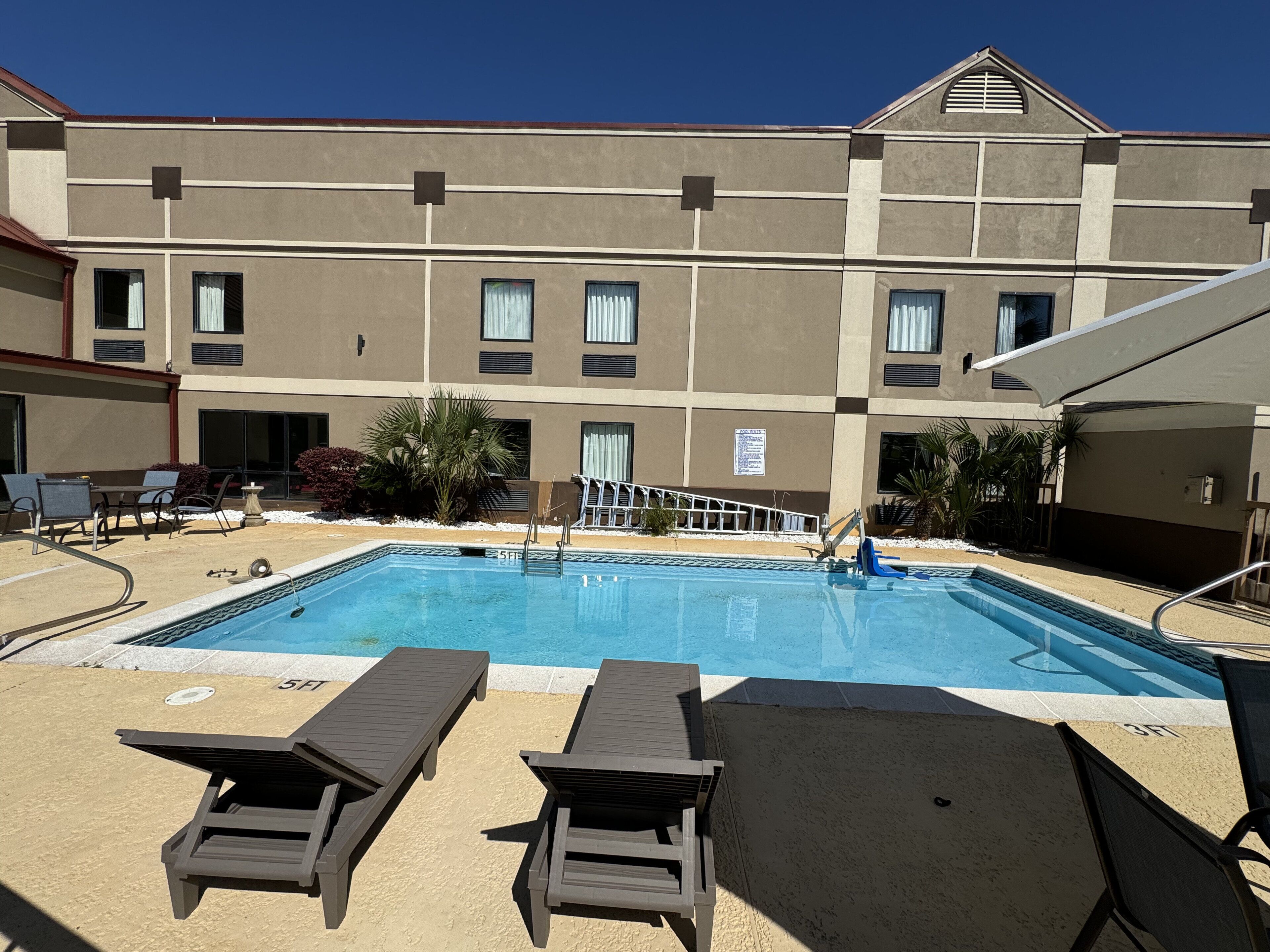 Un área de piscina con sillones de descanso, una piscina y un edificio con múltiples ventanas y un letrero.