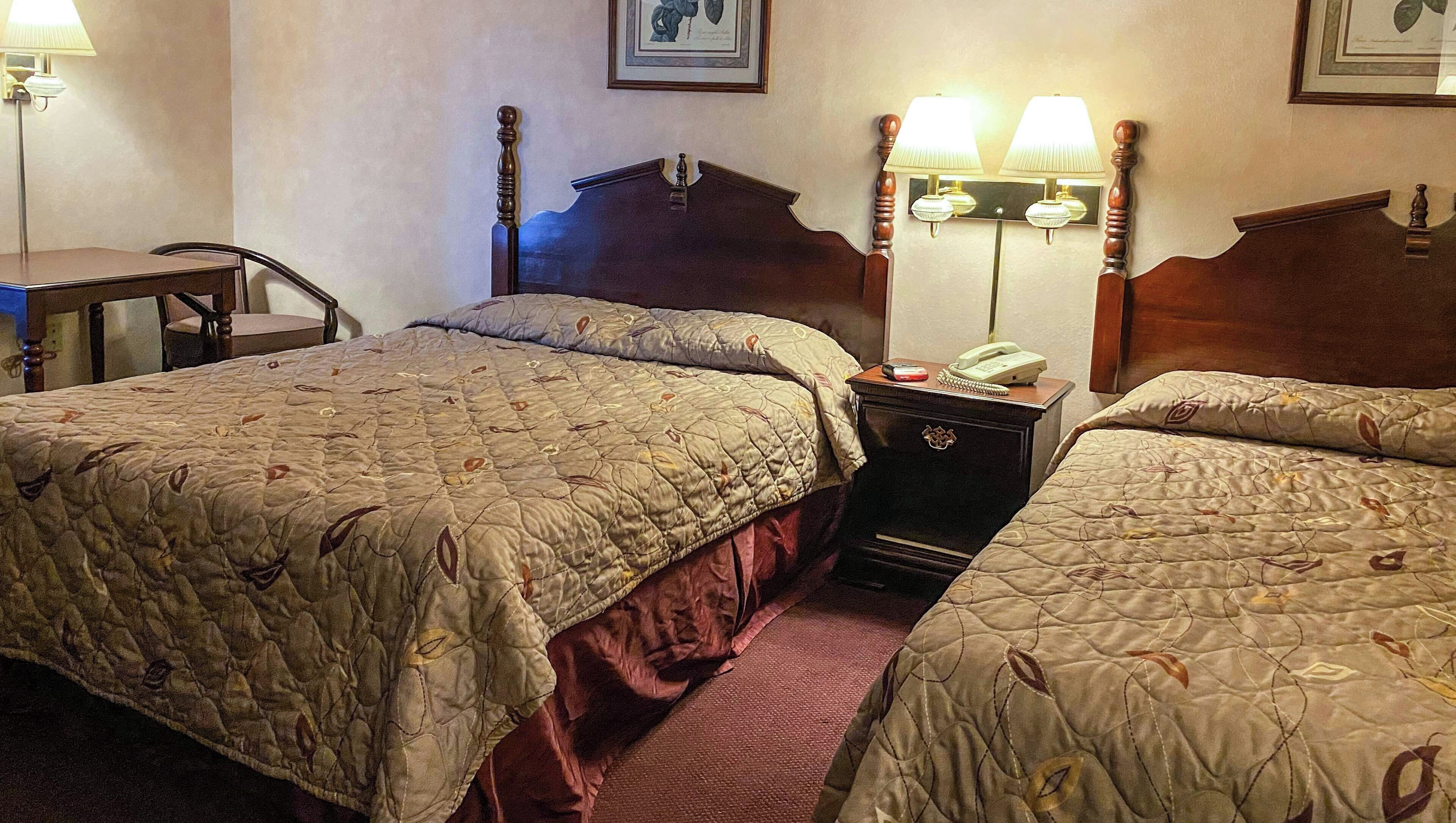 A hotel room with two beds, a desk, a chair, a nightstand with a lamp, and a telephone.
