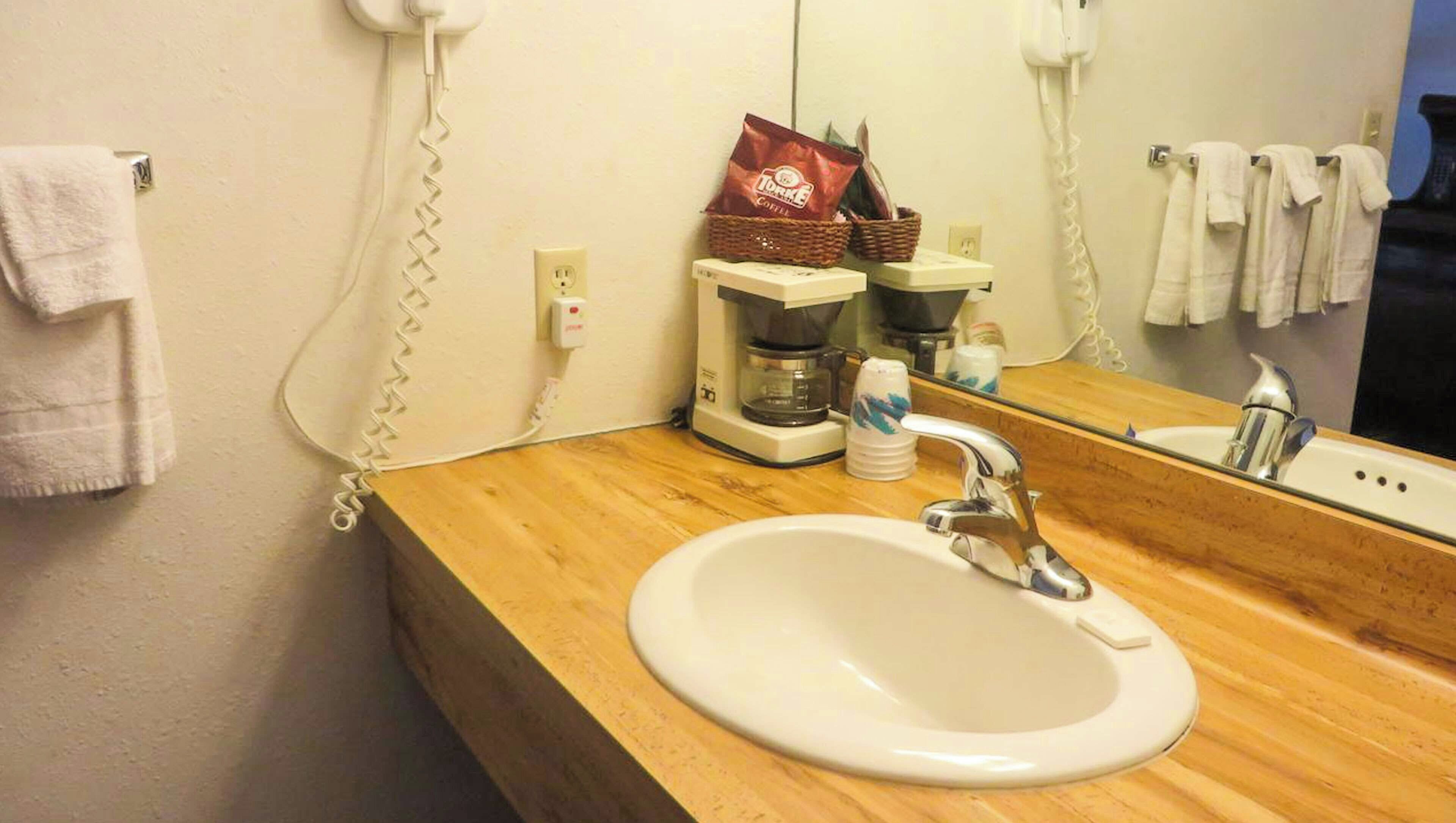 Ett hotellbadrum med en träinredd tvättställsmöbel, en vit handfat, en kaffebryggare, en korg med kaffepåsar och en spegel med en telefon.