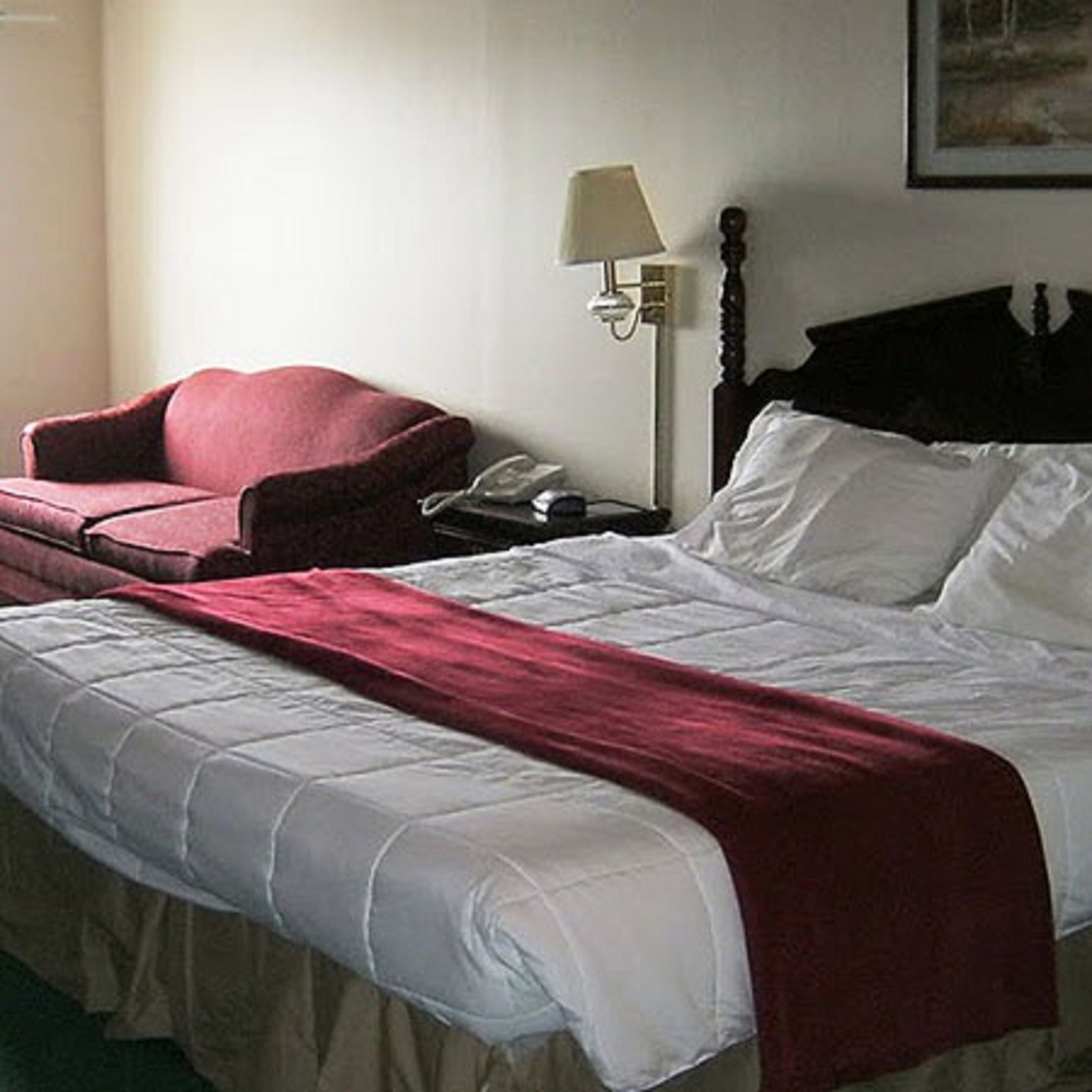 Habitación de hotel con una cama, un sillón rojo, una mesita de noche con una lámpara y un cuadro en la pared.