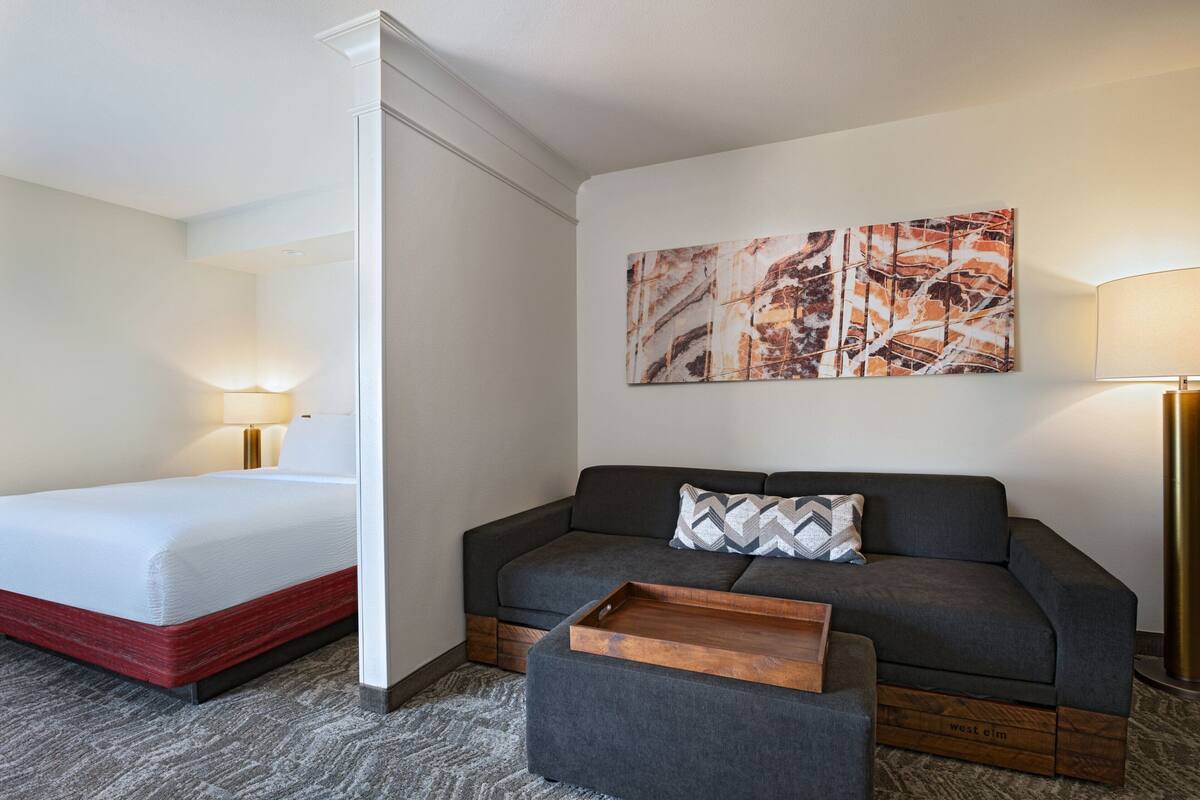 Habitación de hotel con cama, sofá, mesita de madera y un cuadro abstracto colgado en la pared.