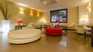 A modern living room with a large round sofa, two armchairs, a red ottoman, and a dining table. There are two lamps, a potted plant, and a window with a view of buildings. - Hotel Don Juan (Granada)