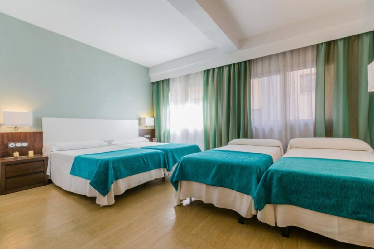 Habitación de hotel con dos camas individuales, una mesita de noche con lámpara y un ventanal con cortinas verdes.