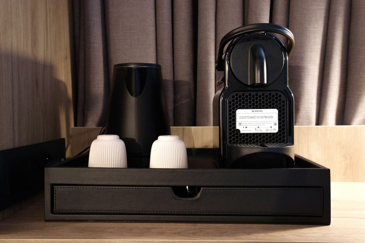 Een zwarte dienblad met een koffiezetapparaat en twee witte flessen, waarschijnlijk koffiemelk of room, voor een houten achtergrond met gordijnen.