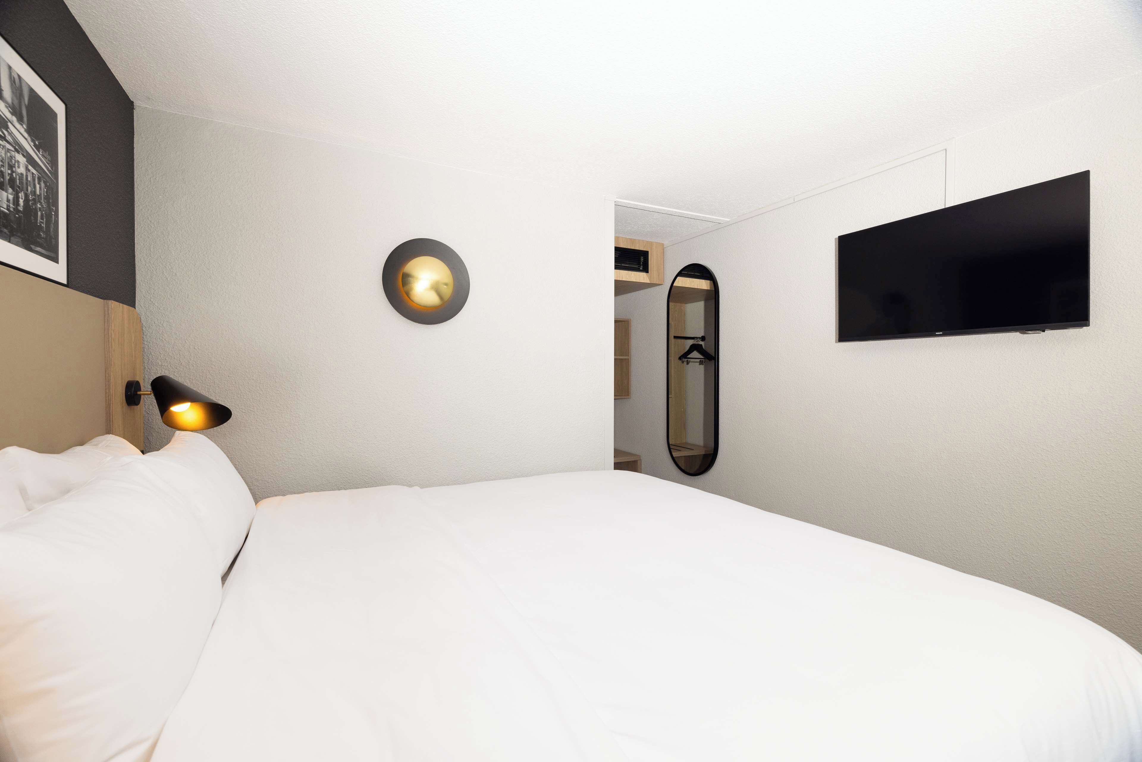 A hotel room with a bed, a wall-mounted TV, a bedside lamp, and a mirror.